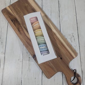 May include: A box of six pastel-colored macarons on a wooden cutting board. The macarons are arranged in a row, with the colors transitioning from pink to blue.
