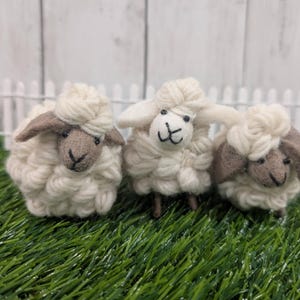 May include: Three handcrafted sheep figures with fluffy white wool bodies and brown faces and ears. The sheep are placed on green artificial grass, in front of a white picket fence.