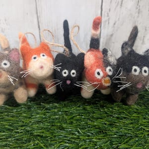 Puede incluir: Cinco adornos de gato de fieltro en varios colores, incluyendo gris, naranja y negro, con bigotes blancos y lazos de cuerda para colgar. Los gatos están sobre un lecho de césped artificial verde.