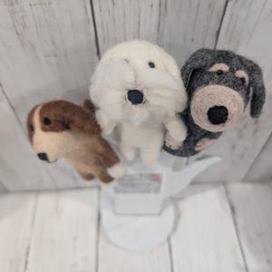 May include: Three felt dog figurines, one white, one brown, and one gray, are arranged in a circle on a white stand. The dogs are all facing the camera.
