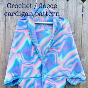 Pode incluir: Um padrão de cardigan de lã polar colorido em tons de azul, rosa e roxo, pendurado em uma cerca de madeira. O cardigan tem um padrão de redemoinho único e é adornado com detalhes de crochê azul claro. O texto "Crochet / fleece cardigan pattern" é visível.