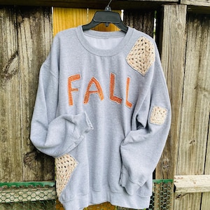 May include: A light gray sweatshirt with the word "FALL" in orange letters across the front. The sweatshirt has beige crochet patches on the sleeves and shoulders. The garment is hanging on a black hanger against a wooden fence.