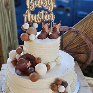 Può includere: Una torta bianca a due piani decorata con palloncini marroni e bianchi, un cappello da cowboy e un paio di stivali da cowboy marroni. La torta è sormontata da un cartello che dice "Baby Austin".
