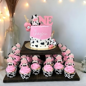 Puede incluir: Un soporte para pasteles de dos niveles con un pastel y cupcakes con temática de vaca rosa y negra. El pastel está decorado con una vaca rosa y blanca, la palabra "ONE" y el nombre "Chaysen". Los cupcakes están cubiertos con glaseado rosa y recortes de vaca.