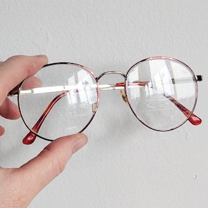 May include: Round eyeglasses with a gold-tone metal frame and red and pink accents. The glasses have clear lenses and red temple tips. The glasses are held up by a hand.