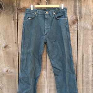 May include: A pair of dark green denim jeans hanging on a wooden hanger against a wood panel background.