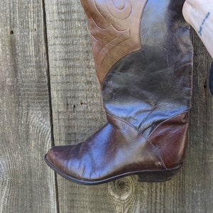 以下が含まれることがあります： 装飾的なパターンがついた、茶色の革製のニーハイブーツ。ブーツはポインテッドトゥで、ローヒールです。