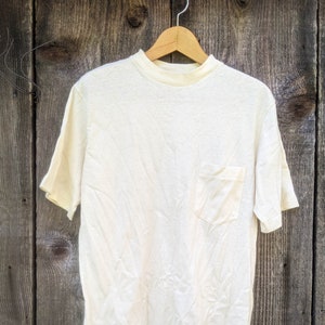 May include: A plain white t-shirt with a single pocket on the left side. The shirt is hanging on a wooden hanger against a woodgrain background.