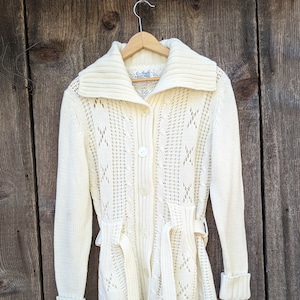 May include: A white knitted cardigan with a button closure, a belt, and a wide collar. The cardigan has a delicate openwork pattern.