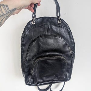 以下が含まれることがあります： 丸みを帯びた形の黒い革製バックパック。上部にハンドル、調節可能なショルダーストラップが2本付いています。バックパックには、前面にジッパー付きポケットと、ジッパー開閉のメインコンパートメントがあります。シルバーカラーの金属製ハードウェアがデザインを際立たせています。