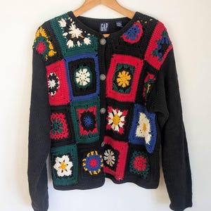 May include: A black cardigan sweater with colorful crochet squares. The squares feature floral and geometric designs in red, yellow, blue, green, and white. The sweater has long sleeves and a button-up front with the GAP logo.