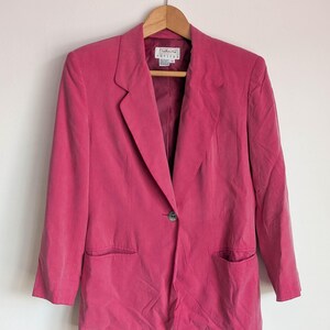 May include: A vibrant magenta blazer featuring a single-button closure, notched lapels, and two front flap pockets. The garment includes a label at the inner neckline that reads Talbots Petite. It is displayed on a wooden hanger against a plain wall.