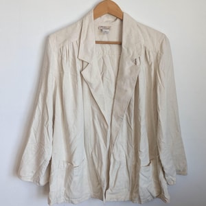 Puede incluir: Blazer color crema con solapa de muesca y dos bolsillos delanteros, colgado de una percha de madera. La chaqueta tiene un corte holgado y mangas largas. La tela parece ser una mezcla de lino o algodón.