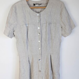 May include: A light beige linen dress with short sleeves, a round neckline, and a button-down front. The dress features two chest pockets and pleats at the waist. It is hanging on a wooden hanger against a white background.