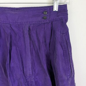 May include: A close-up of a vibrant purple skirt hanging on a hanger. The skirt features a high waist with two dark buttons, pleats, and a pocket detail. The fabric has a slightly wrinkled texture.