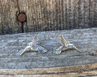 May include: A pair of silver hummingbird earrings. The hummingbirds are facing opposite directions and have detailed wings.