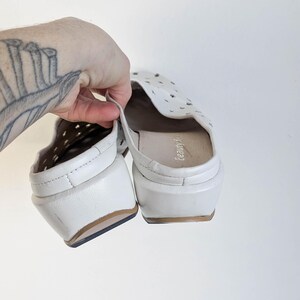 May include: White leather slip-on shoes with a perforated design on the top. The shoes have a small heel and a brown sole. The interior of the shoe is a light tan color. The shoes are held up by a hand.
