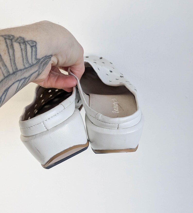 May include: White leather slip-on shoes with a perforated design on the top. The shoes have a small heel and a brown sole. The interior of the shoe is a light tan color. The shoes are held up by a hand.