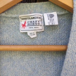 May include: A light blue wool sweater with a Crazy Horse label. The label features a red horse logo and the text "SHETLANDER CRAZY HORSE 100% WOOL". The sweater is hanging on a wooden hanger.