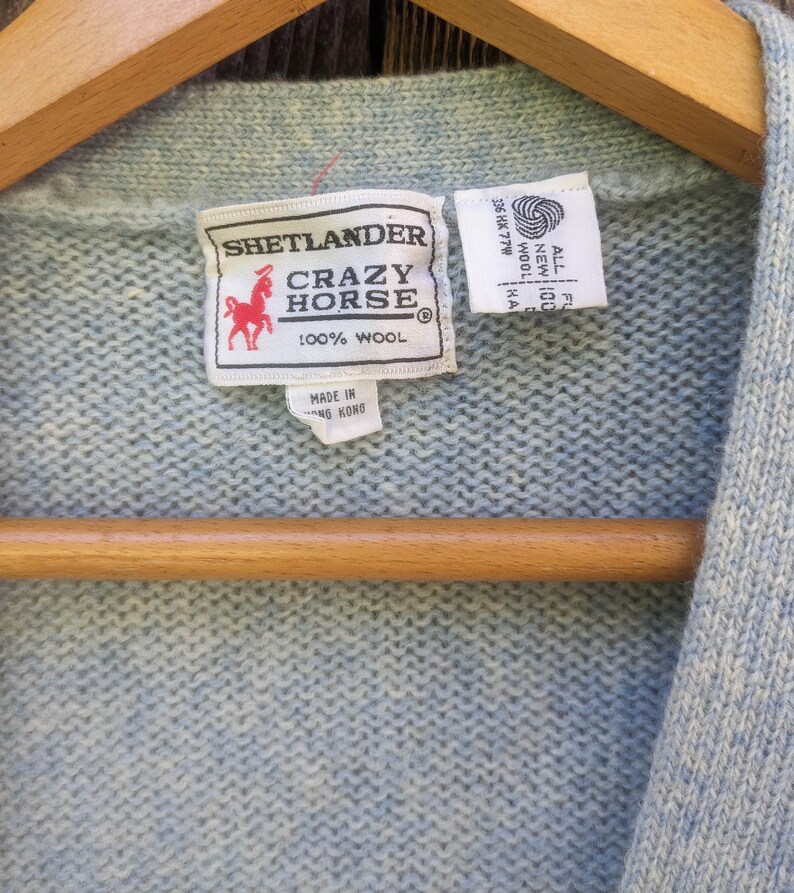 May include: A light blue wool sweater with a Crazy Horse label. The label features a red horse logo and the text "SHETLANDER CRAZY HORSE 100% WOOL". The sweater is hanging on a wooden hanger.