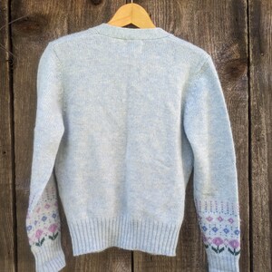 May include: A light blue cardigan sweater with a ribbed hem and cuffs. The sleeves feature a floral pattern in pink, blue, and green. The sweater is hanging on a wooden hanger against a weathered wood background.