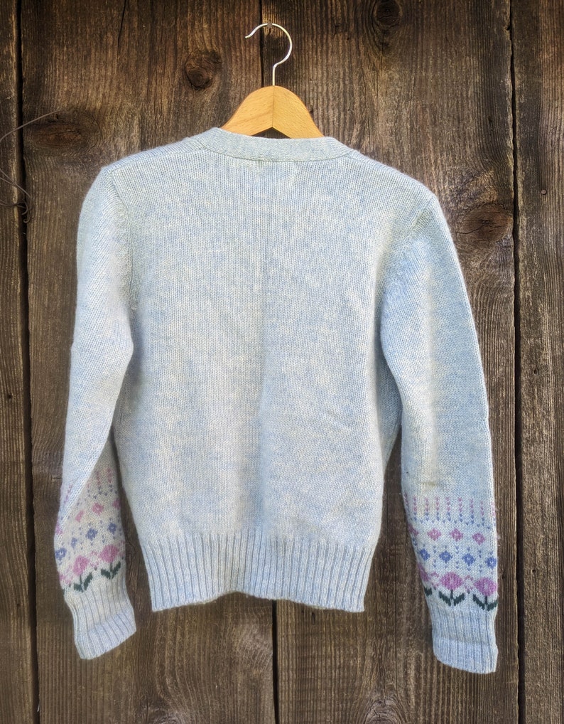 May include: A light blue cardigan sweater with a ribbed hem and cuffs. The sleeves feature a floral pattern in pink, blue, and green. The sweater is hanging on a wooden hanger against a weathered wood background.