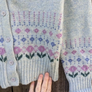 May include: Light blue cardigan sweater with a floral pattern in shades of purple, green, and blue. The sweater has a button-up front and ribbed cuffs and hem. The design features diamond and flower motifs.