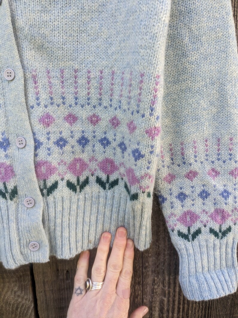 May include: Light blue cardigan sweater with a floral pattern in shades of purple, green, and blue. The sweater has a button-up front and ribbed cuffs and hem. The design features diamond and flower motifs.