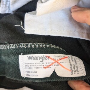 May include: A dark green pair of Wrangler jeans with a white care label. The label says "Machine wash warm with like colours, only non-chlorine bleach when needed, tumble dry low. Made in USA, 100% Cotton."