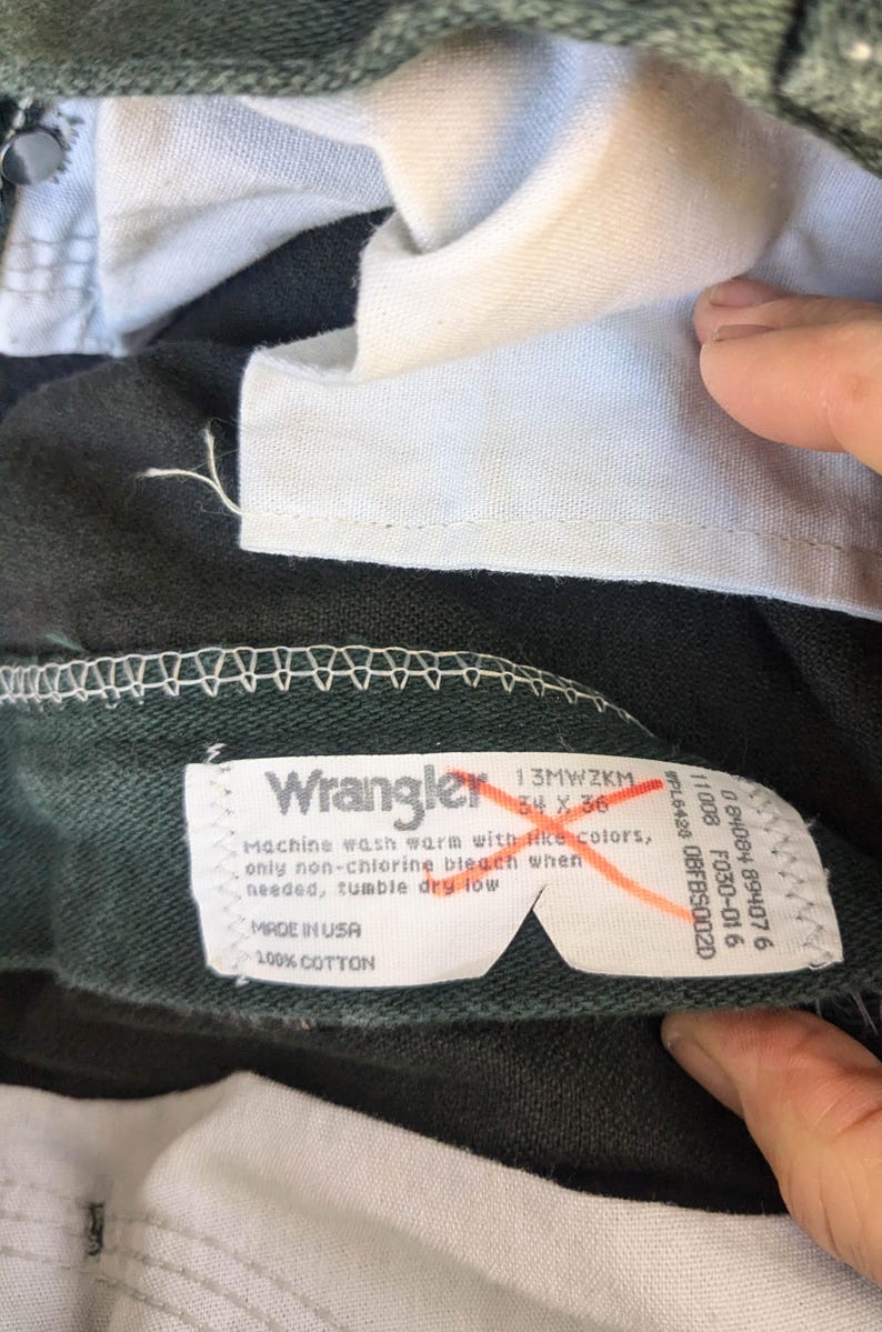 May include: A dark green pair of Wrangler jeans with a white care label. The label says "Machine wash warm with like colours, only non-chlorine bleach when needed, tumble dry low. Made in USA, 100% Cotton."