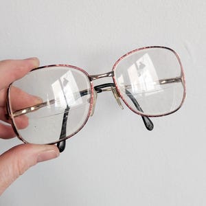May include: Vintage eyeglasses with large, rectangular lenses. The frames are a combination of gold-tone metal and a patterned red and brown material. The temples are black, and the glasses are held by a hand against a white background.