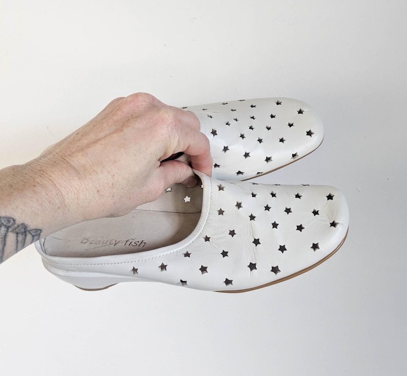 May include: White leather slip-on shoes with star-shaped cutouts. The shoes have a rounded toe and a low heel. The brand name "beauty fish" is visible inside one shoe. The shoes are shown against a white background.
