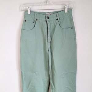 Puede incluir: Vaqueros de cintura alta verde menta con el texto "LIFE TIME" encima del botón. Los vaqueros tienen el estilo clásico de cinco bolsillos y cuelgan de una percha de plástico transparente. Los pantalones son de color sólido.