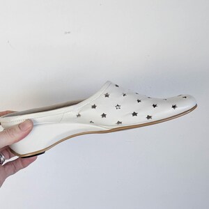 May include: White leather slip-on shoe with star cutouts. The shoe has a wedge heel and a brown sole. The shoe is held by a hand.