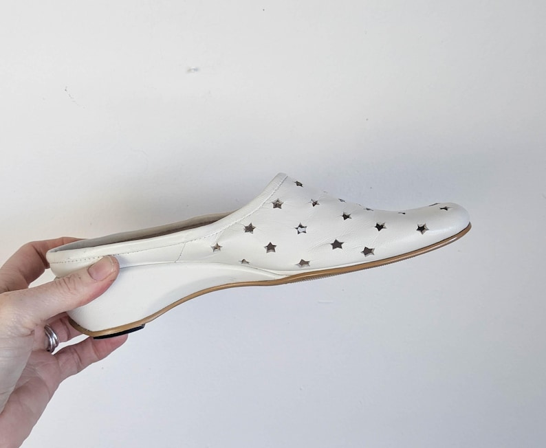 May include: White leather slip-on shoe with star cutouts. The shoe has a wedge heel and a brown sole. The shoe is held by a hand.