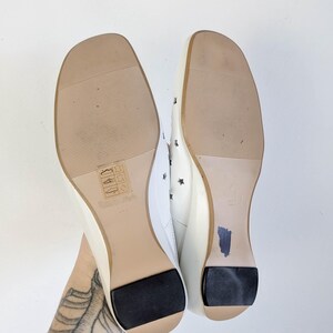 May include: Pair of white flats with square toes, featuring star cutouts and black heel pads. The soles are light brown with a textured pattern. The size is marked as 23.