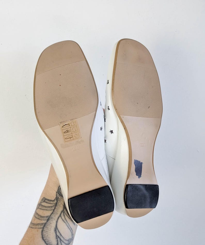 May include: Pair of white flats with square toes, featuring star cutouts and black heel pads. The soles are light brown with a textured pattern. The size is marked as 23.