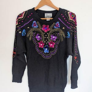 Pode incluir: Camisola preta com um design floral colorido feito de lantejoulas e contas. A camisola tem um decote redondo e mangas compridas. O rótulo diz "Mamie West".