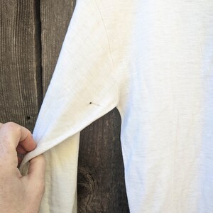 May include: White long-sleeved shirt with a slight texture. The shirt is hanging on a wooden surface.