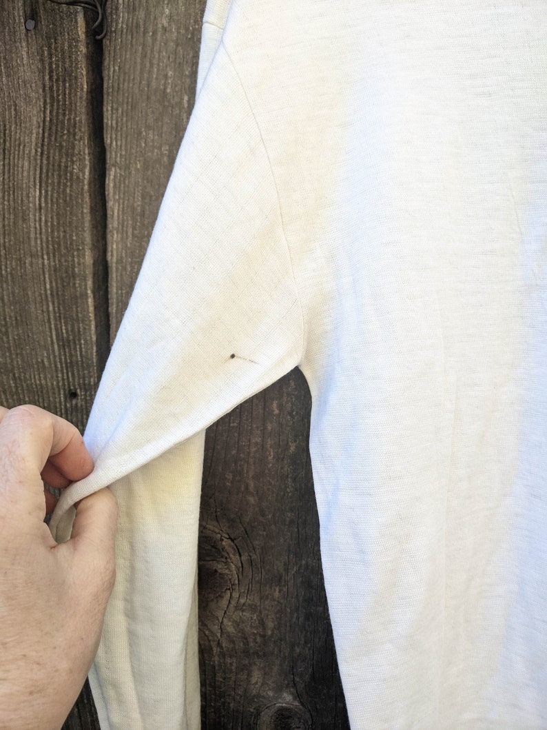 May include: White long-sleeved shirt with a slight texture. The shirt is hanging on a wooden surface.