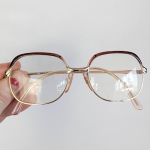 May include: Vintage eyeglasses with a gold-tone metal frame and clear lenses. The top of the frames are a dark brown color. The glasses are held by a hand against a white background.