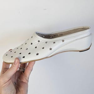 May include: White wedge heel shoe with star cutouts. The shoe has a rounded toe and a small wedge heel. The sole is a light brown color. The shoe has the numbers "9015 230" printed on the inside.