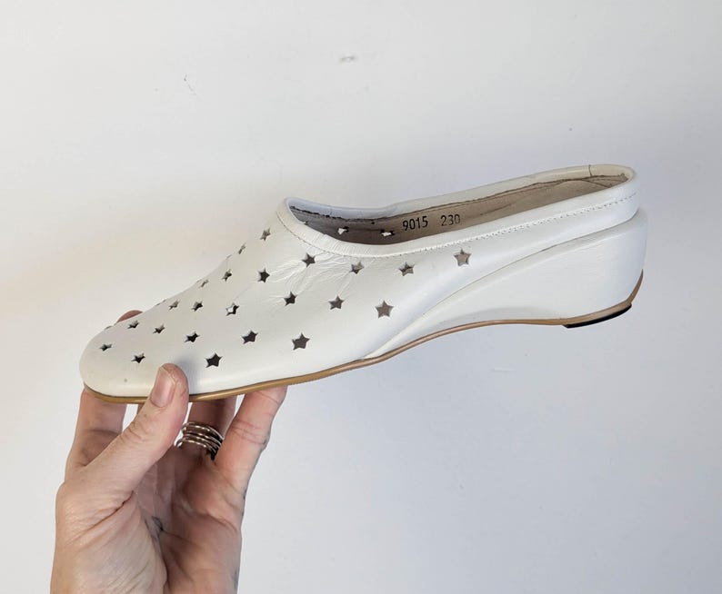 May include: White wedge heel shoe with star cutouts. The shoe has a rounded toe and a small wedge heel. The sole is a light brown color. The shoe has the numbers "9015 230" printed on the inside.