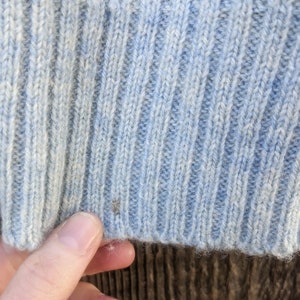 May include: Close-up of a light blue knit sweater with a ribbed texture. The sweater has a subtle pattern in a darker shade of blue and green at the top. The bottom edge of the sweater is visible, showing the knit construction.