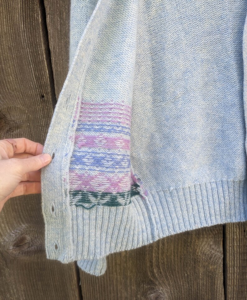 May include: Light blue cardigan with a ribbed hem and button closure. The inside panel features a colorful geometric pattern in shades of purple, blue, and green. The cardigan is made of a soft, textured knit fabric.