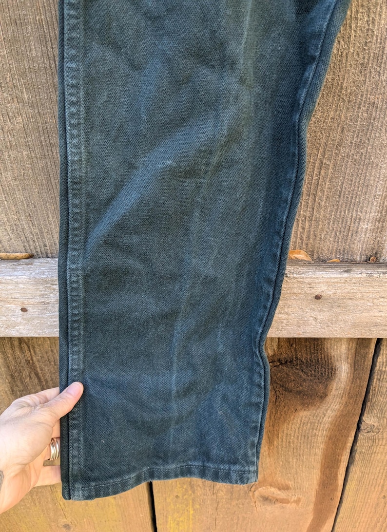 May include: Close-up of the dark green fabric of a pair of trousers. The fabric has a subtle texture and a slight sheen.