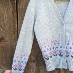 May include: Light blue cardigan sweater with a floral pattern in pink, purple, and blue. The sweater has a V-neck, button closure, and long sleeves. The label reads "Shetland Crazy Horse".