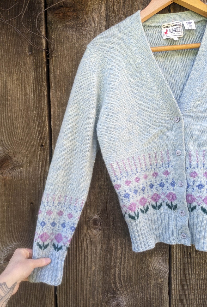 May include: Light blue cardigan sweater with a floral pattern in pink, purple, and blue. The sweater has a V-neck, button closure, and long sleeves. The label reads "Shetland Crazy Horse".