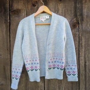 May include: A light blue cardigan sweater with a floral pattern in pink, purple, and green. The sweater is button-down and hangs on a wooden hanger against a wooden background. The floral pattern is on the bottom of the sweater and sleeves.