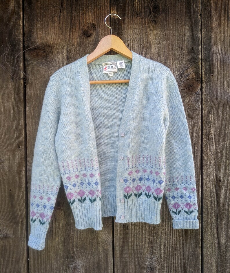 May include: A light blue cardigan sweater with a floral pattern in pink, purple, and green. The sweater is button-down and hangs on a wooden hanger against a wooden background. The floral pattern is on the bottom of the sweater and sleeves.